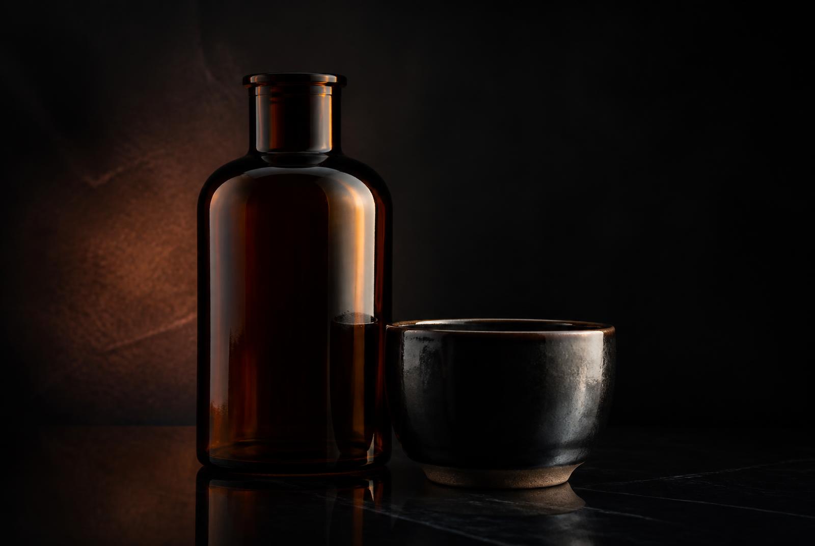 Produktaufnahme einer unbedruckten Glasflasche mit Keramikschale im dunklen Studio